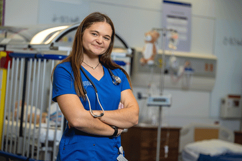 A female nursing student poses for the camera with a nursing lab in the background.